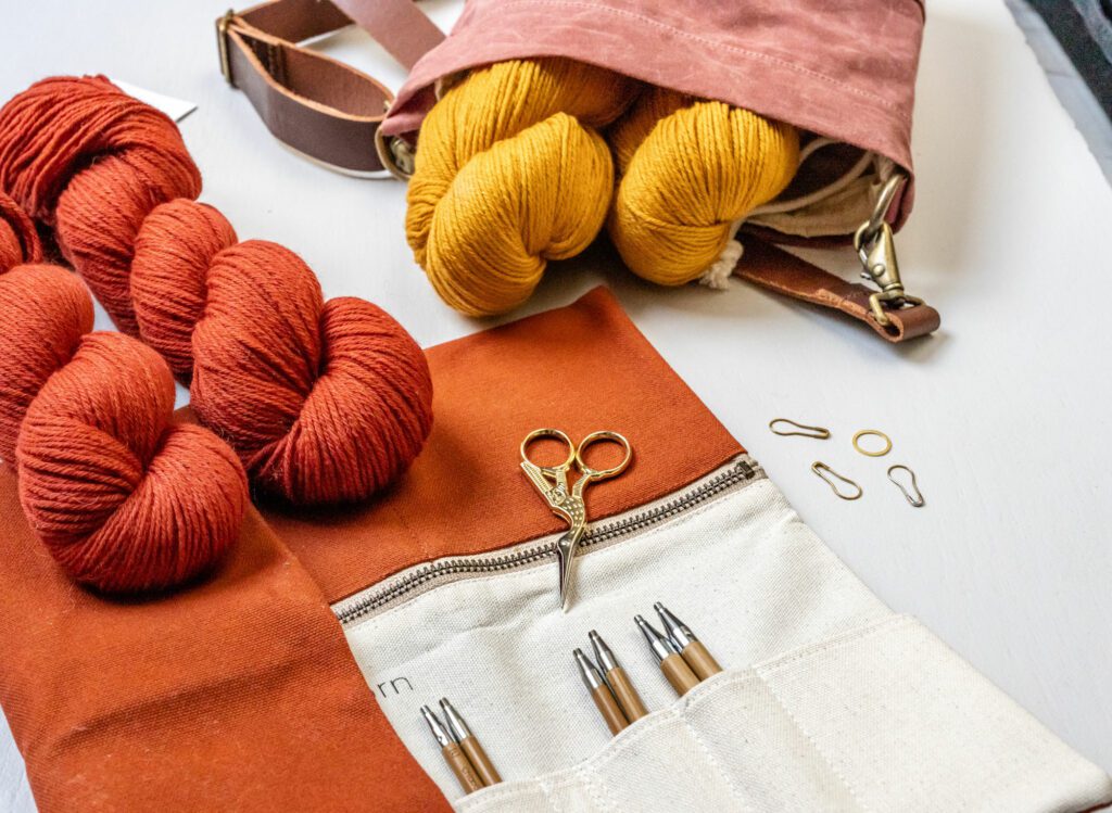 Zwei rote und zwei curry farbene Stränge von Rosy Green Wool liegen auf dem Tisch.  Dazu sieht man ein aufgeklapptes Nadeletui von Twig and Horn sowie die Canvas Bucket Bag.